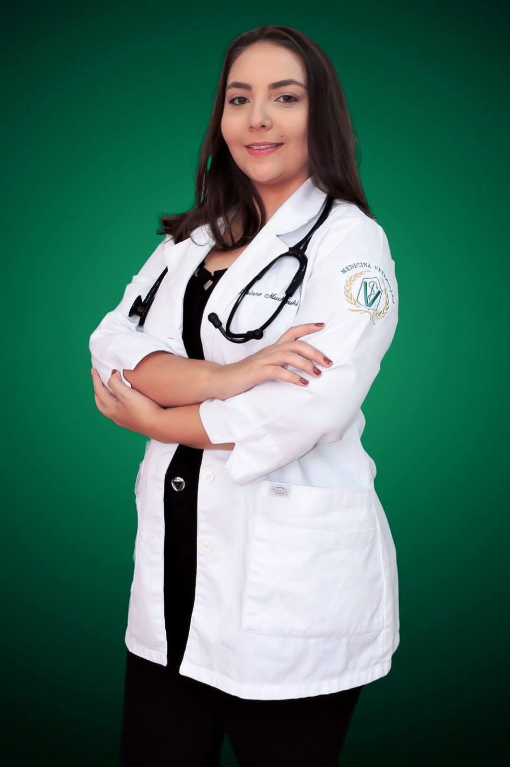 Gianinne, médica-veterinária, de jaleco branco, estetoscópio e braços cruzados diante de um fundo verde